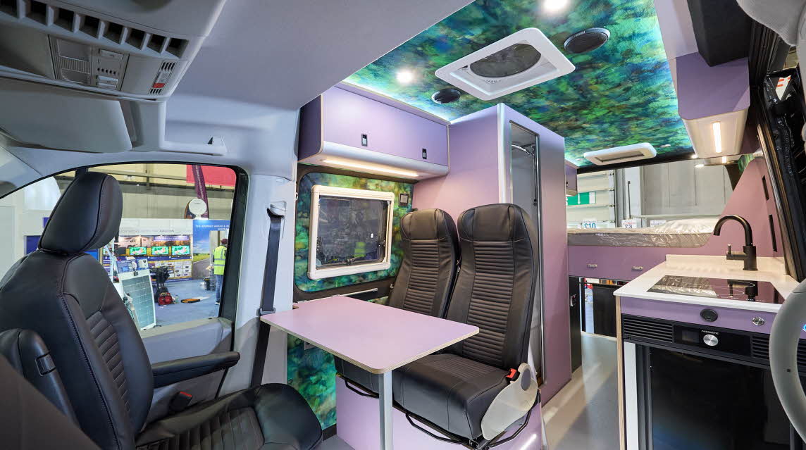 Vanworx MaxTraxx Crafter’s interior has black leather upholstery.  There is a white table and its furniture is white.  It has a patterned internal roof.  At the rear there is a fixed bed.