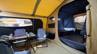 The Trigano Camplair S has yellow coloured canvas with a cream room divider.  The bedroom door is open.  There is a step to the bedrooms.  There is a light on the ceiling.  There is a dining table with four blue chairs