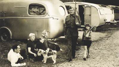 Family sitting in front of caravans in Elstree as smiling Home Guard soldier and small boy march past together