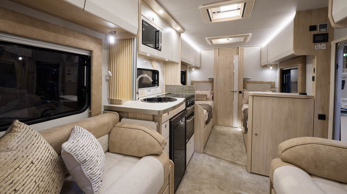 The Elddis Avalon 285 has beige carpets and cream cabinets.  The upholstery is cream and pale brown.  The kitchen is placed in the middle and just behind there are two fixed single beds.  The open interior door leads to the washroom.