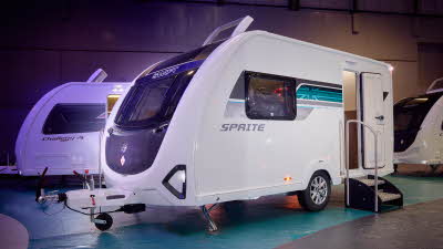 Swift Sprite Alpine 2’s exterior is white with a black panelled front.  There is a turquoise decal to the right of the side window with the Union flag.  The entrance door is open and there are two metal steps to gain easy access