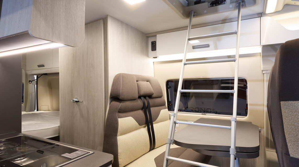 The Roller Team Livingstone 2’s upholstery is dark cream and dark brown.  There is a grey table in front of the sofas.  The cabinets are pale wood.  There is a ladder leading to the open pop top roof.