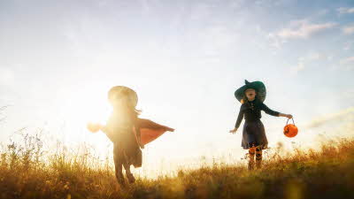 Two young children dressed up as witches for Halloween run across the grass with the setting sun behind them