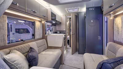 Swift Challenger Exclusive 560’s interior has wooden furniture with dark locker doors and beige upholstery which has embossed patterns.  There are three cushions on each sofa.  The rear door is open revealing the bedroom