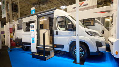 The Sun Living V Series 65SL is white with brown and bronze decals.  There are two steps outside the open sliding door.  There is an interactive stand next to the steps and the rear door is open.