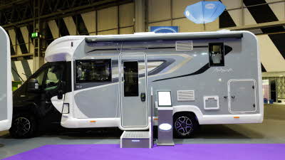 The Bailey Autograph 72-2 has a black cab and grey coachbuilt body.  It has black, cream and grey decals.  There is a step outside the habitation door with an interactive stand next to it.  Above it is a blue Bailey umbrella.