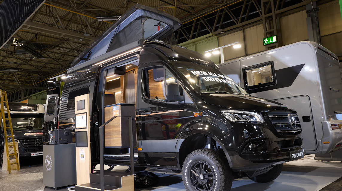 The Adria SuperTwin has a black exterior.  The habitation door is open and there are two steps to gain easy access. There is an information board to the left of the steps.  The rising roof is open and so are the rear doors.