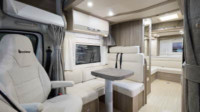 Benimar Primero 282’s interior has cream upholstery.  The furniture is wooden.  The rear seats have seat belts.  The table can be extended and is folded.  There are tied up cab window curtains.  There is a rear lounge.  