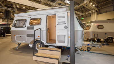 The classically styled Eriba Touring 530 Urban has a two tone layout in silver and white.  Its pop top roof is open as is its entrance door.  There are two wooden steps to gain easy access.