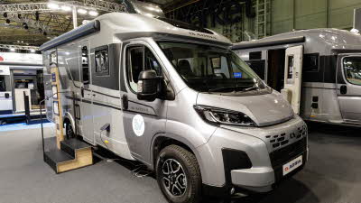 The Adria Compact Supreme DL is silver with a white coachbuilt body.  It has dark grey decals along its side.  There are two steps up to the main door to enable easy access.  It has an interactive stand by the side of the steps.