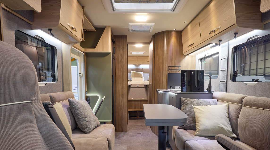 The Eura Mobil Integra Line GT 726 EF has beige carpets and wooden cabinets.  The upholstery is mid brown.  There is a grey table (folded) in front of the sofas.  There are two steps to the rear fixed bed.