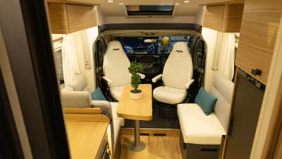 The Pilote P740FC Evidence has cream upholstery complemented with wood furniture.  The front two seats are swivelled to face inside the van.  There is a wooden table which is shown as folded.  There's a turquoise cushions on each sofa.
