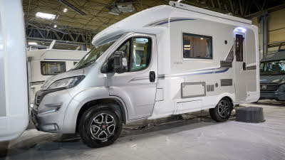 Auto-Sleeper Nuevo EK Plus' exterior has a silver cab with a white body and grey/blue decals.  The entrance door is closed and there is a step to gain easy access.  