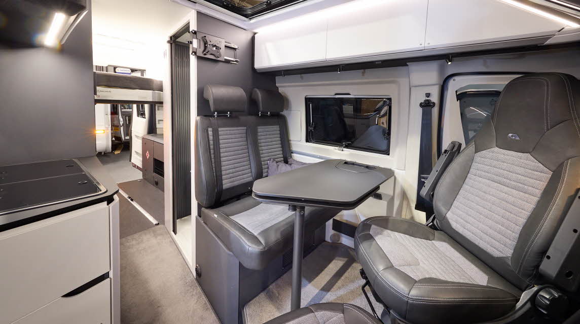 The Adria Twin Supreme’s floor has a pale brown carpet, with white cabinets and dark grey panels.  There is a large panoramic sunroof.  The upholstery is two tone with dark edging.  There is a grey table which can be rotated.  