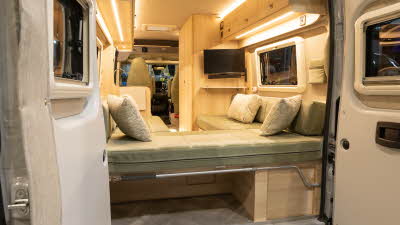 The Vantage NEO has a green u shaped sofa with patterned cushions.  The cabinets are made of pale wood.  Beneath the sofa there is a storage area.  Both rear doors are open.