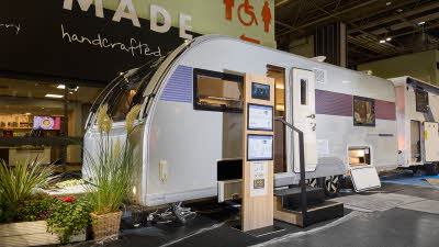 The Adria Alpina Colorado has a silver exterior with a blue decal that turns to purple at the rear, its entrance door is open and there are two wooden steps to gain easy access. 