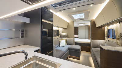 Interior of the Adria Alpina Rio Grande with an unusual front kitchen, grey sofas, cosy cushions and wood accents, illuminated by natural light through windows and skylights, offering a view outside.  Perfect for relaxation and designed for comfort.