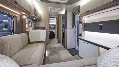 Interior of the Swift Conqueror Grande 650L with beige sofas, cosy cushions and beige carpets.   The large windows and four sky lights will let natural light flood in.  The door to the rear bedroom is open.