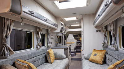 Auto-Sleeper Warwick XL has light grey embossed upholstery, with cream and grey overhead cabinets.  It has light wooden panelling, there are two gold cushions on each sofa and each window has two patterned curtains