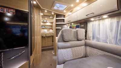 The Carthago C2-tourer I 148 RB-QB has a wooden floor, with cream cabinets. The upholstery is grey with white stitching. There is a step up to the rear fixed bed.