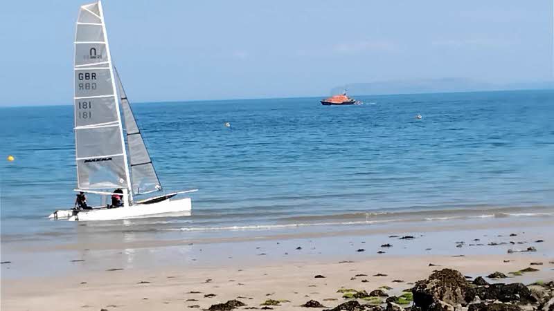 A view from the the beach at Traeth Bychan, Anglesey. A small sailboat near the shore on the shallow water. An RNLI boat is in the distance on the darker blue deeper waters. The sky is a very pale blue.
