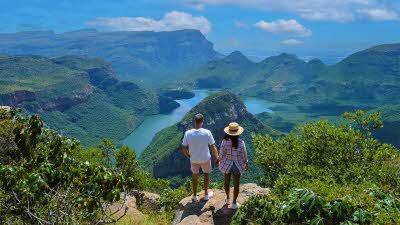 Two people standing on rocks looking over the stunning the Blyde river canyon with lush greens blue river and pale blue sky with fluffy clouds