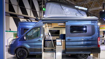 Bailey Endeavour B64's exterior is blue with black decals, and the roof is raised.  There are two steps to gain easy access and there is an interactive plinth next to them.  The sliding door is open revealing the lounge.