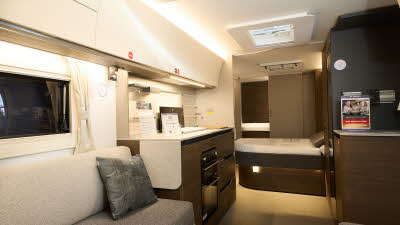 Adria Alpina Mississippi’s interior, it’s cream with a large panoramic window.  The upholstery is two tone grey.  The fixed bed is beyond the kitchen with the rear washroom’s door is open.