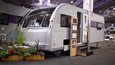 The Adria Adora 623 DP Tiber’s exterior has light grey walls with a patterned decal from its open door to the rear.  There are two wooden steps with an interactive display to the left.  There is an open locker.  There is a floral display.