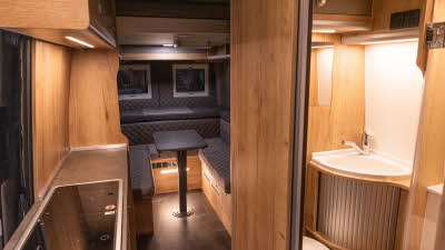 The Mega Mobil Mega Lounge 680 S Twin has grey upholstery with wooden walls and cabinets.  The kitchen is in the bottom left with the washroom opposite showing a basin with a cupboard with a Tambour door.