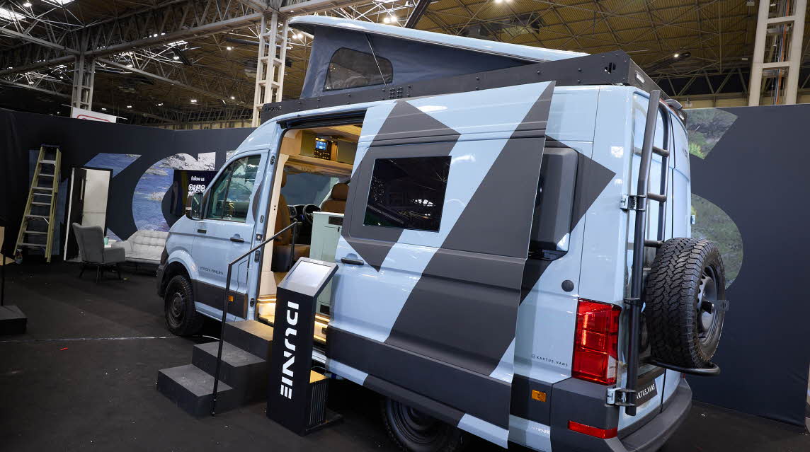 The Kaktus Adventure Vans Dune x4 has a pale blue and dark grey exterior with prominent decals.  There are two steps beside the open sliding door.  The pop top roof is fully extended.