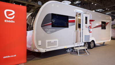 The Elddis Crusader Borealis has a grey exterior with grey, red and black decals.  The entrance door is open and there is a metal step to gain easy access.  There is a large Elddis red banner with their website address next to it.
