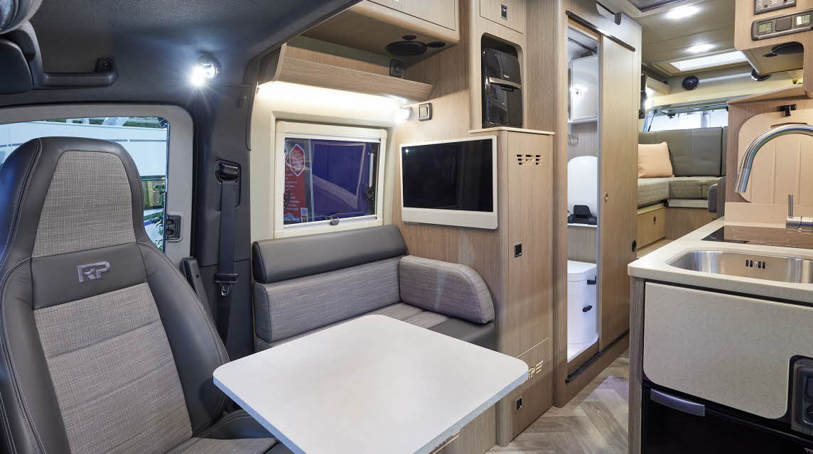 RP Motorhomes Rebellion 2 has two tone grey upholstery.  There are light wooden overhead lockers and door to the washroom.  On the floor is wooden laminate.  There is a folding flap which gives additional kitchen work space.