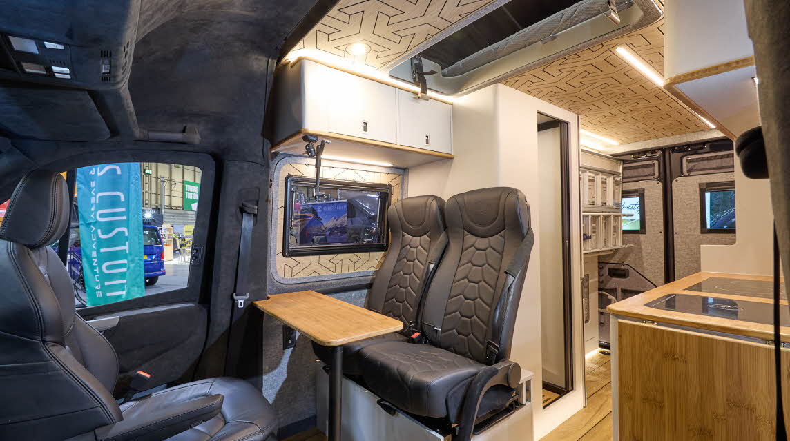 Knights Custom Conversion Prestige XL has wooden flooring with black upholstery.  The cabinets are white and pale wood.  The washroom door is open.  The pop top roof is open.
