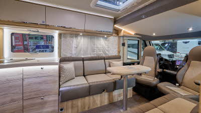 Eura Mobil Integra Line IL’s interior has brown and beige upholstery.  Its furniture is a mixture of pale and dark wood with cream doors.  The table can be extended and is showing folded in half.  The front two seats are swivelled.