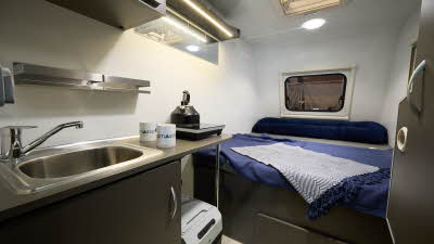 Campmaster Duo GLX interior, kitchen sink and mixer tap, with two mug and a kettle, fixed double bed with blue duvet cover