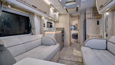 Elddis Crusader Zephyr’s interior has grey upholstery with dark grey accents.  It has pale wooden furniture.  The kitchen is in the middle and the bedroom door is open revealing a fixed island transverse bed.