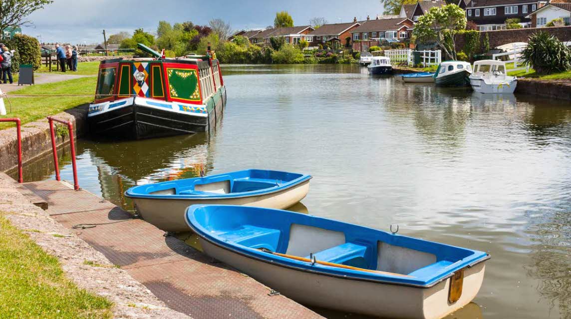 two small blue rowing boats on a perfectly still canal with a brightly painted narrowboat moored up behind them