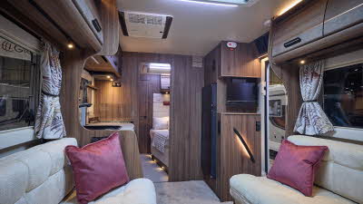 The Auto-Sleeper Burford has a pale grey carpet, with dark wood cabinets. There is a roof light. The upholstery is cream with two burgundy cushions. There is a fixed bed to the rear.