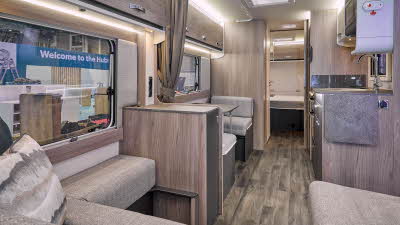 Swift Sprite Exclusive Quattro MB’s interior has a beige U shaped sofa with wooden furniture, there is a dinette opposite the kitchen unit.  The rear door is open revealing the fixed island bed.  