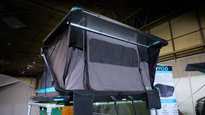 The SkyPod Lair is displayed on a silver frame.  It has grey canvas walls and a black hard roof.  It has turquoise flashes around it.  There is a black ladder.  