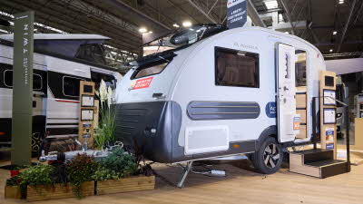 The Adria Action 361's exterior has light grey walls with dark blue bumpers and trim. The front window and panoramic sunroof are both open. The entrance door is open with two wooden steps.  There is a floral display around its hitch