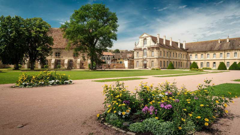 View of the old abbey of Cluny in France. With flowers in the foreground with a gravel path running alongside the fresh green grass with the abbey behind under a blue sky