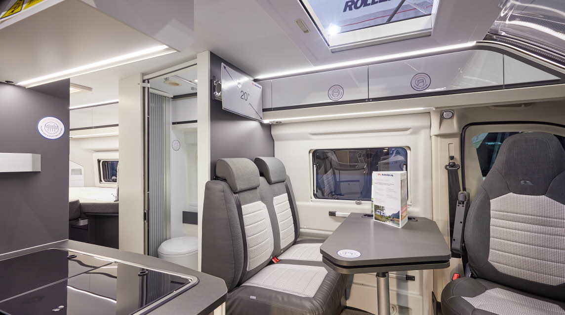 Adria Twin Supreme 640 SLB interior, rear passenger seats in brown and cream, fixed table, swivelled driver’s seat which can double as a dining chair.  Also shown is a hob, an open roof light and the combined washroom and toilet.  
