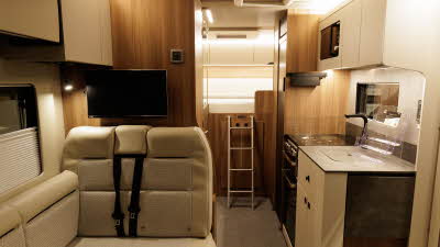 The The Auto-Trail Imala 736 G has beige patterned upholstery.  The overhead lockers match.  There are wooden walls.  To the rear there are fixed beds which can be accessed via the silver ladder.