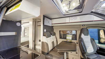 Adria Twin Sports 600 SPB interior, rear passenger seats in brown and cream, fixed table, swivelled driver’s seat which can double as a dining chair.  Also shown is a hob, access to the rising roof and the combined washroom and toilet.  