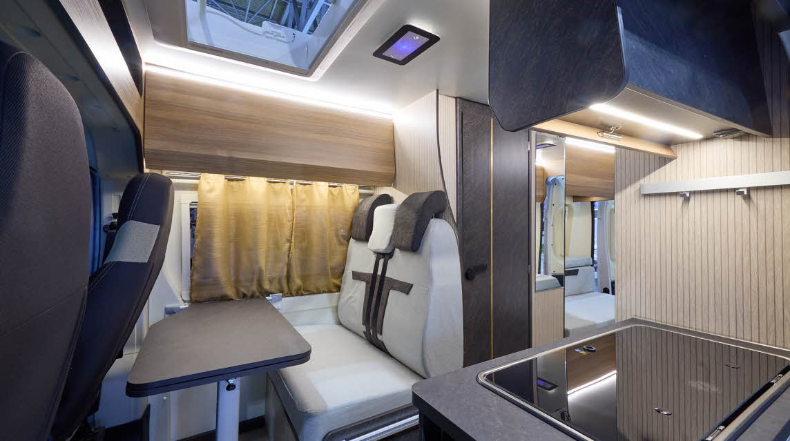 The Rimor Horus 40 has a wooden floor.  The upholstery is grey and dark brown.  The cabinets are all pale wood.  The skylight is open.  The covered hob and sink are at the front.  There is a rear lounge and the rear door is open.