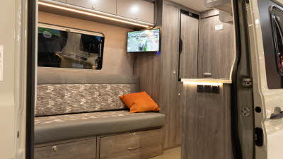 The Rolling Homes Camper Ltd Darwin FL (MWB) has grey/brown upholstery with wooden cabinets.  There is a TV below one of the overhead lockers.  There is an orange cushion on the sofa/