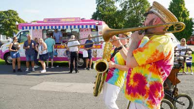 Musicians in tie-dye shirts march past a pink ice cream van