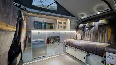 Auto-Sleeper Air's interior has two tone beige upholstery with gold inserts.  The upholstery has gold piping.  The kitchen including appliances are silver.  The rising roof is open.
